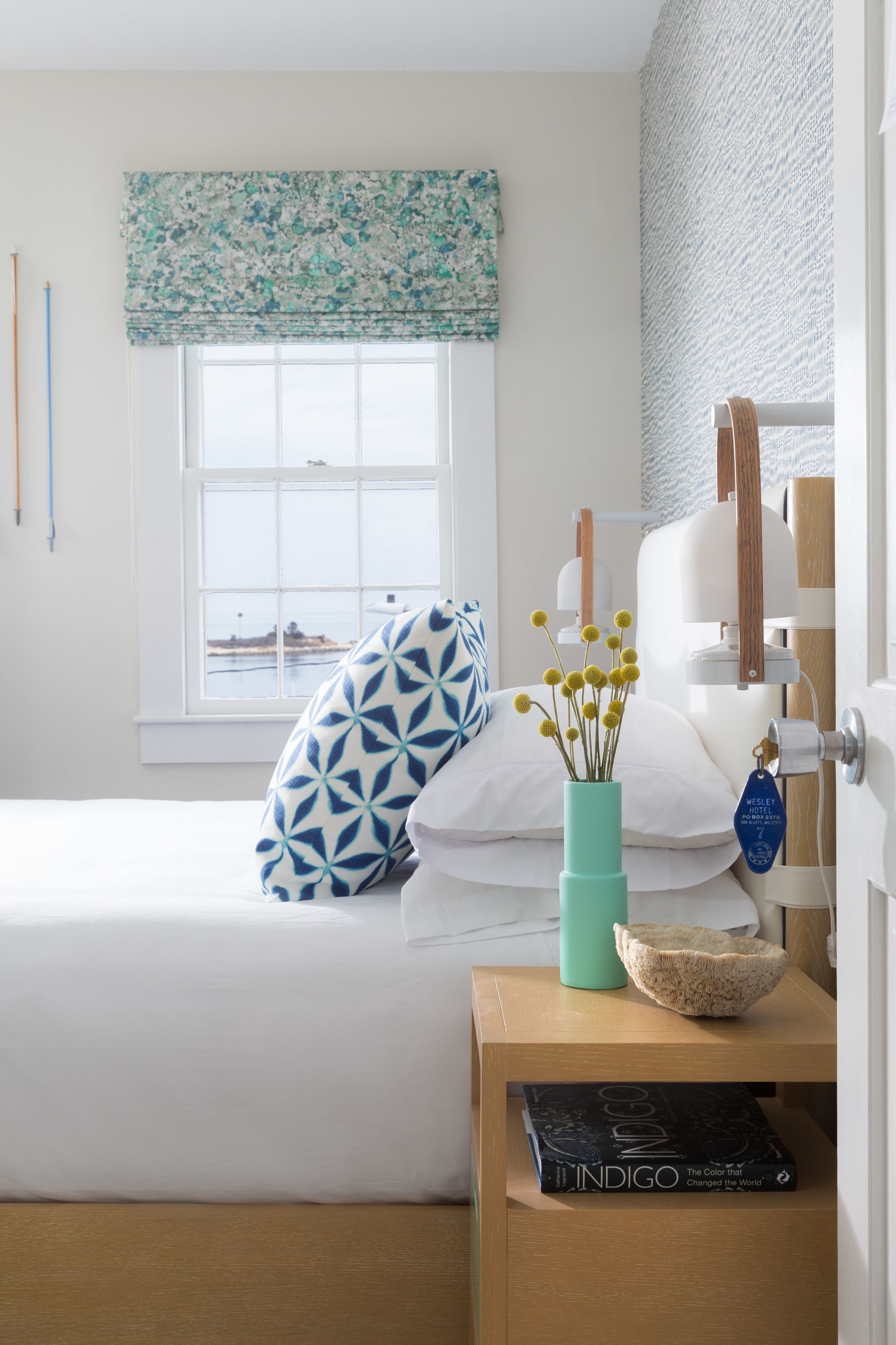Un dormitorio ordenado con cama, una mesita de noche con un libro, un florero con flores y una ventana con vista al mar.