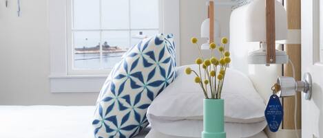 Una camera da letto ordinata con un letto, un comodino con un libro, un vaso con fiori e una finestra vista mare.