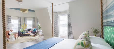 Quarto de hotel moderno com uma cama grande, área de estar com sofá e cadeiras, e vista da cidade através de grandes janelas.