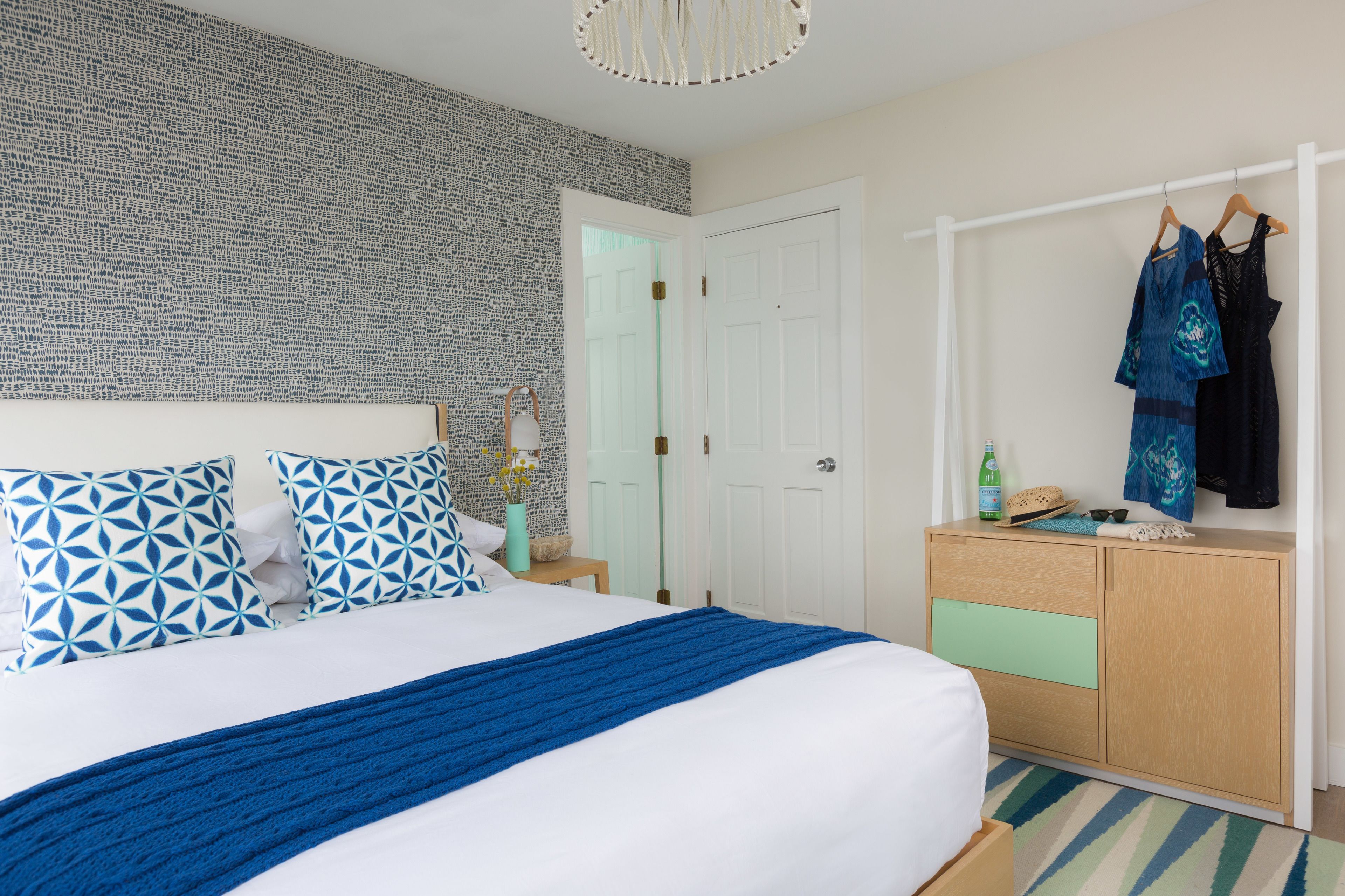 Una habitación de hotel ordenada con un papel tapiz decorativo, una cama con ropa de cama azul y blanca, un tocador de madera y un perchero con dos vestidos.