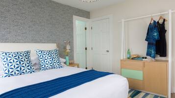 Una habitación de hotel ordenada con un papel tapiz decorativo, una cama con ropa de cama azul y blanca, un tocador de madera y un perchero con dos vestidos.