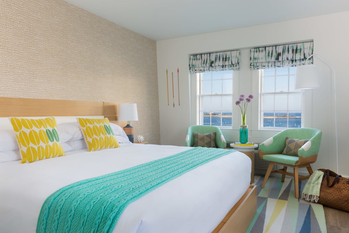 A hotel room with a large bed, a green armchair, a small table with a vase of flowers, and a view of the sea through the window.