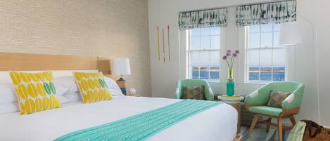 A hotel room with a large bed, a green armchair, a small table with a vase of flowers, and a view of the sea through the window.