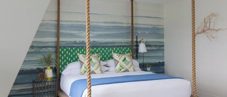 Un lit à baldaquin avec vue sur la mer par une fenêtre, une petite table de chevet avec une lampe et un élément décoratif mural.