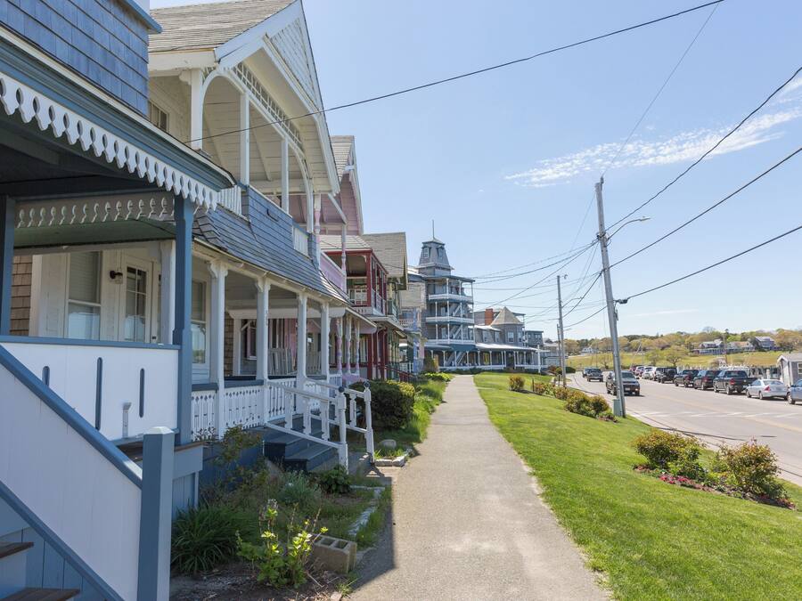 A row of traditional houses with front porches, a sidewalk, and a street with parked cars.