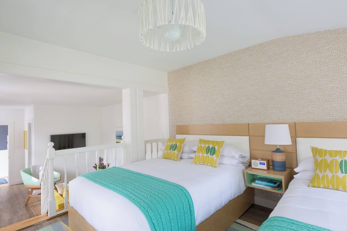 A hotel room with two beds, a television, a bedside table with a lamp, and a small shelf with books.