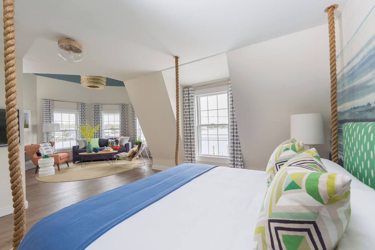 A spacious room with a large bed, a seating area with a sofa and armchairs, and a view of the outdoors through large windows.