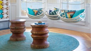 A room with a hanging light fixture, two white wicker chairs with colorful cushions, a wooden table, and a view of the water through large windows. - Summercamp (Oak Bluffs)