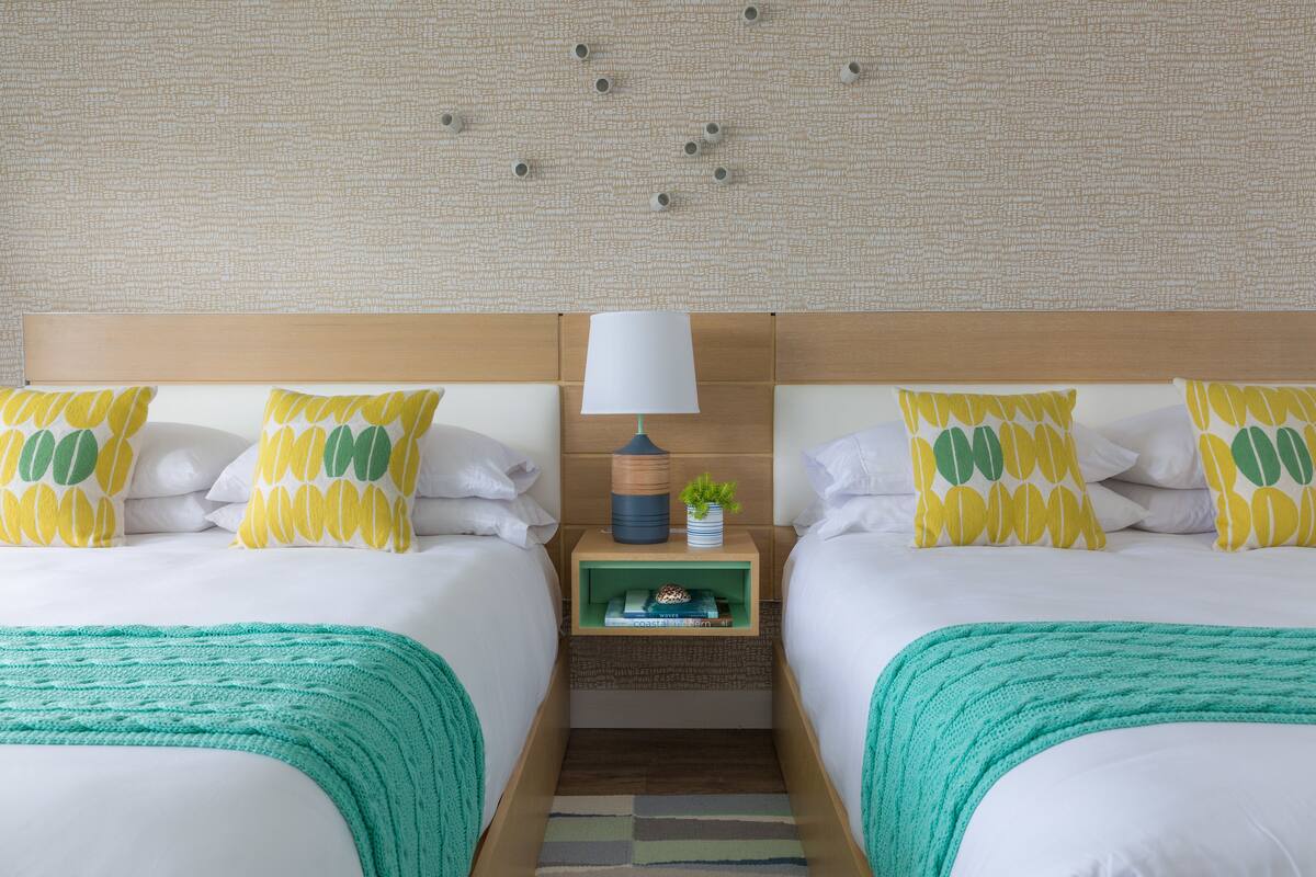 A hotel room with two beds, a nightstand with a lamp, and a small shelf with books and a plant.