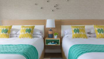 A hotel room with two beds, a nightstand with a lamp, and a small shelf with books and a plant.