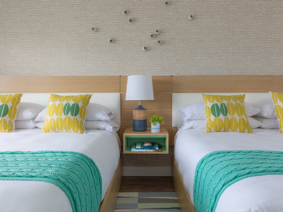 A hotel room with two beds, a nightstand with a lamp, and a small shelf with books and a plant.
