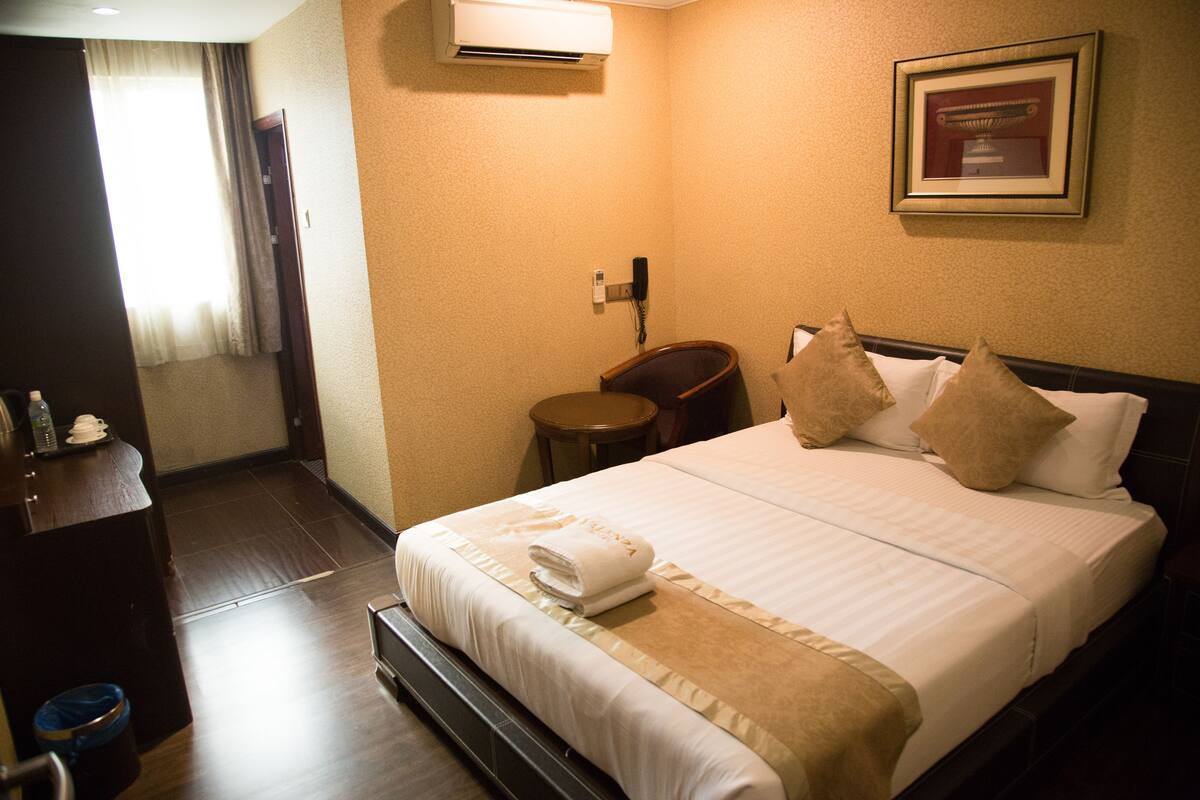 A hotel room with a neatly made bed, a chair, a small table, and an air conditioning unit on the wall.