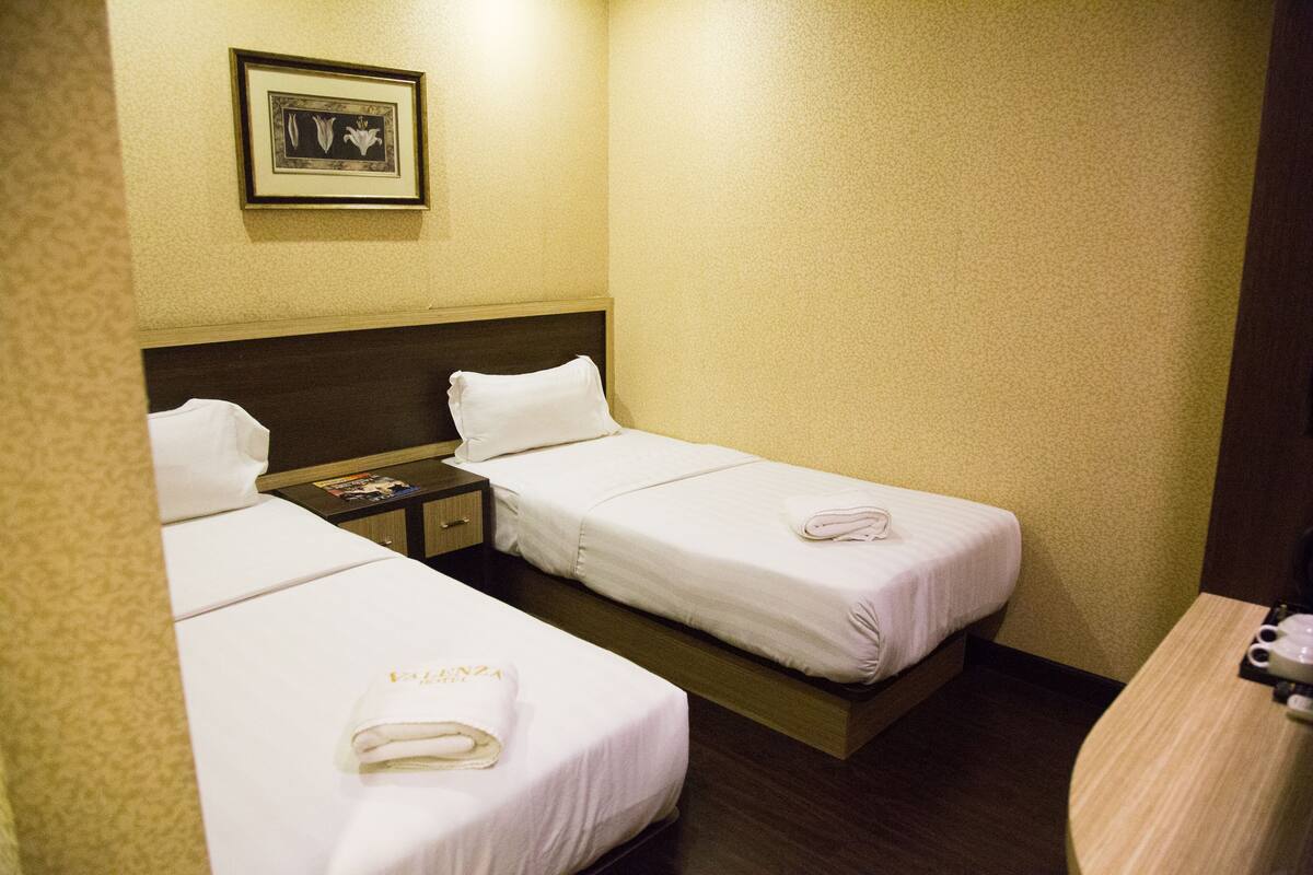 A hotel room with two single beds, a nightstand with a book and a framed picture, and a wooden desk with a cup and saucer.