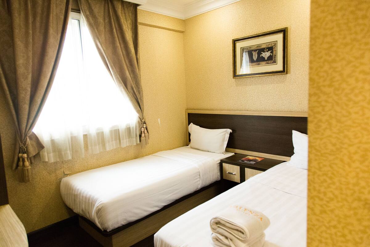 A hotel room with two single beds, a nightstand, a framed picture on the wall, and a window with curtains.