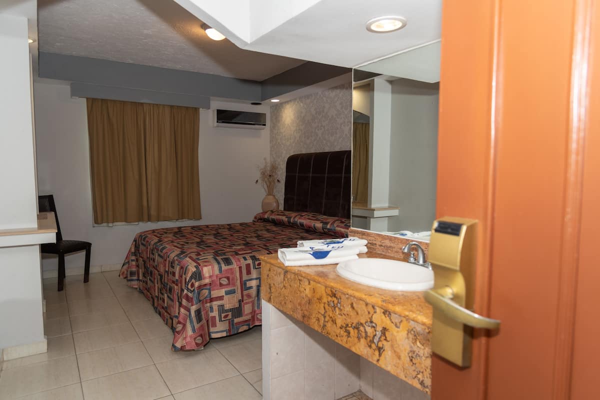 Une chambre d’hôtel avec un lit, une chaise, une salle de bain avec un lavabo et un miroir, et une porte.