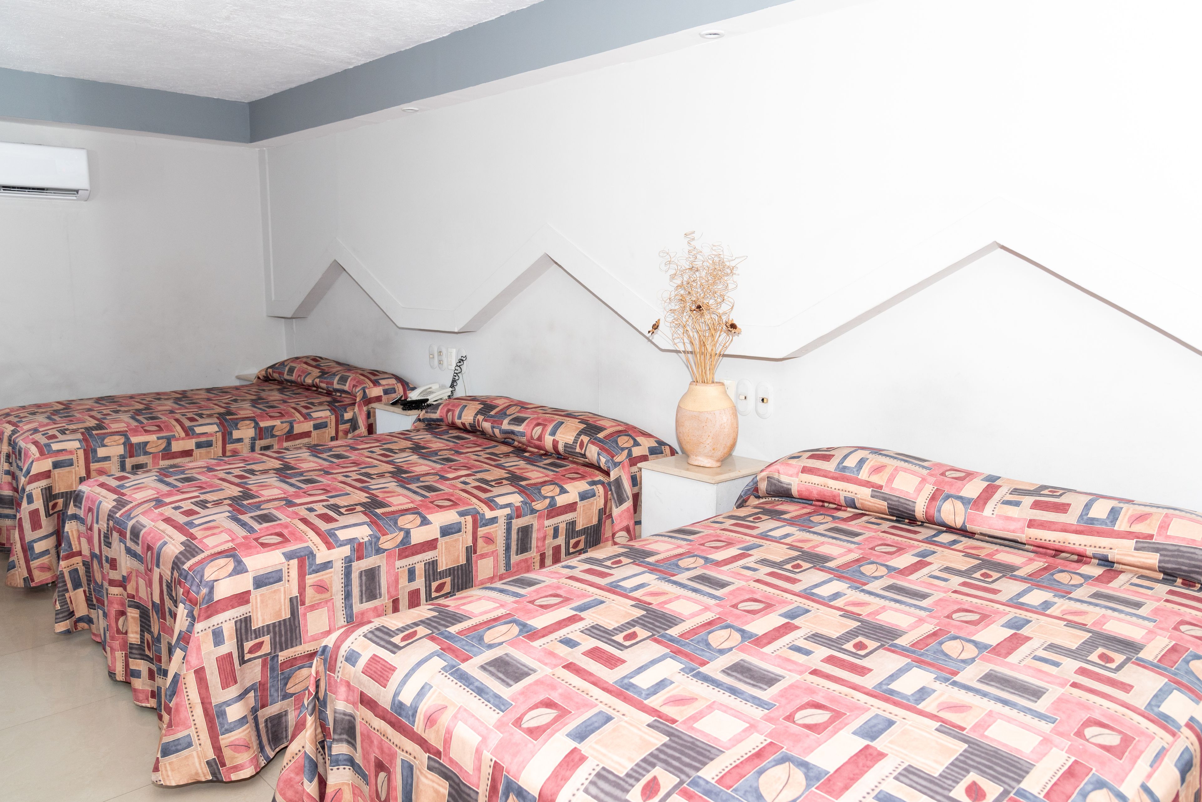 A hotel room with three beds, each with patterned bedding, a vase with dried flowers on a nightstand, and a white air conditioning unit on the wall.