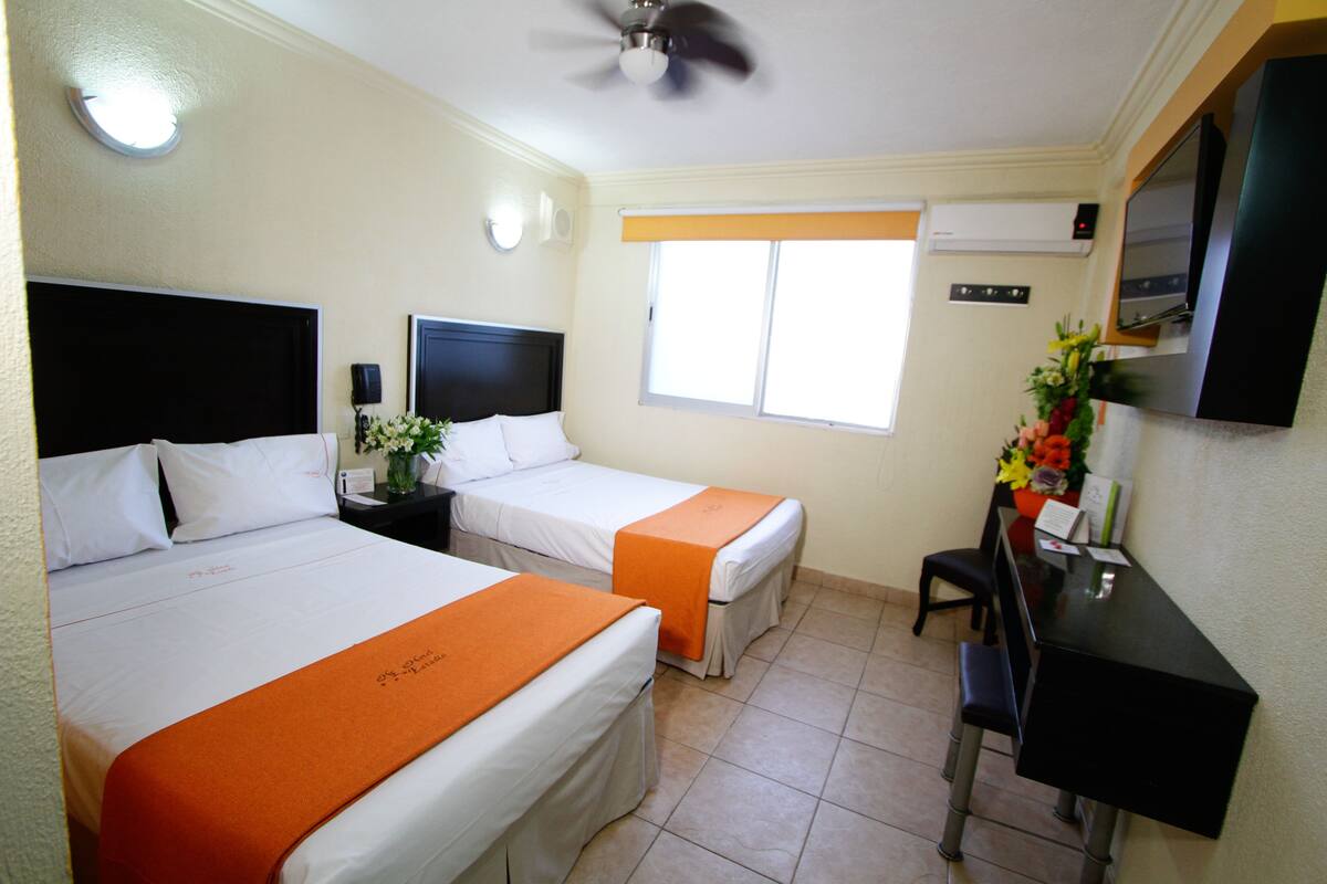 A hotel room with two beds, a desk, a chair, a ceiling fan, a window, and a TV on the wall.