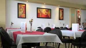 Une salle à manger avec des tables recouvertes de nappes blanches, des chaises noires et des sets de table rouges. Les murs sont ornés d’œuvres d’art encadrées et un présentoir à menus se trouve au centre de la pièce.
