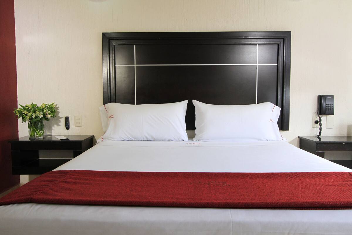 A hotel room with a large bed, a red throw blanket, a nightstand with a plant, and a phone on the nightstand.