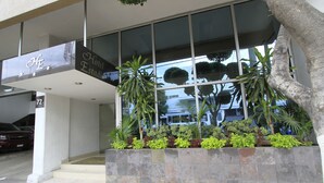 L’entrée d’un hôtel, avec une enseigne indiquant « Hôtel Estadio », un chemin bordé de carreaux et un jardinier planté de différentes espèces.
