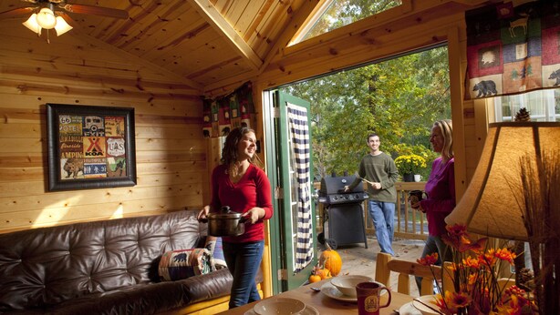 Een gezellig houten interieur met een houten plafond, een eettafel met borden en een mok, een leren bank en een groot raam met uitzicht op bomen.