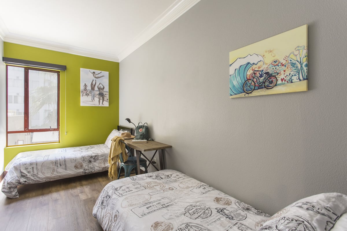 A room with two beds, a table with a backpack, a bicycle, and a window with a view.