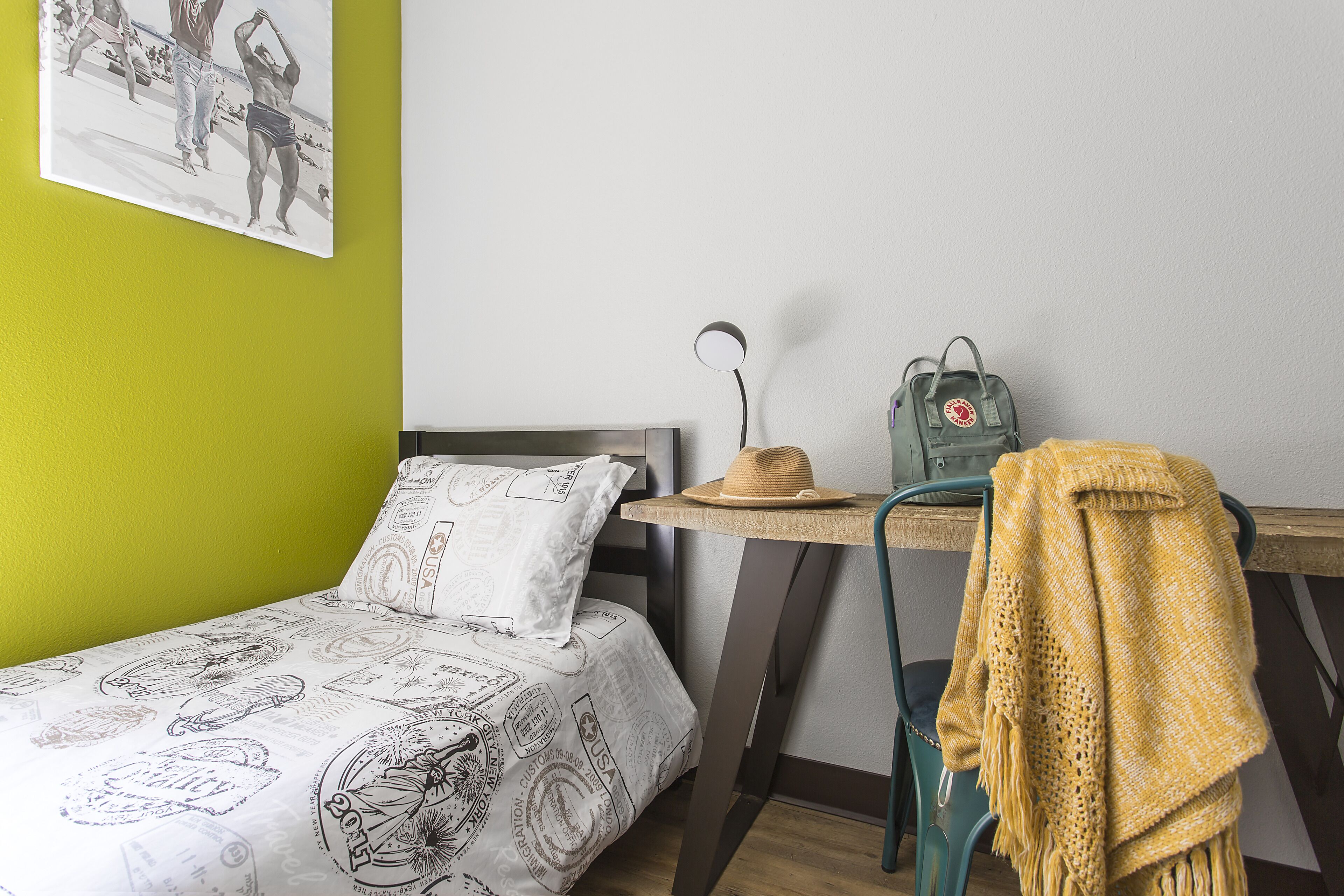 A room with a bed, a desk, a chair, a backpack, and a wall with a picture.