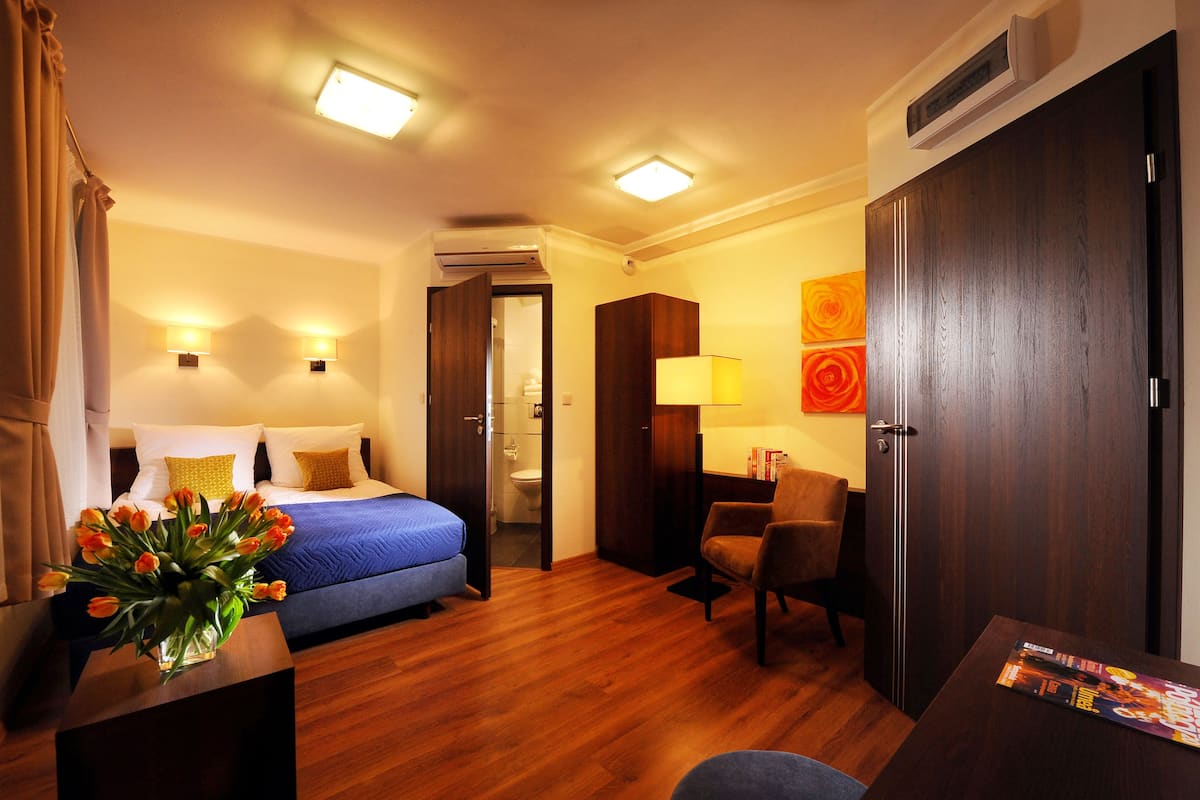 A hotel room with a bed, a desk, a chair, a vase of flowers, and a wall-mounted air conditioner.