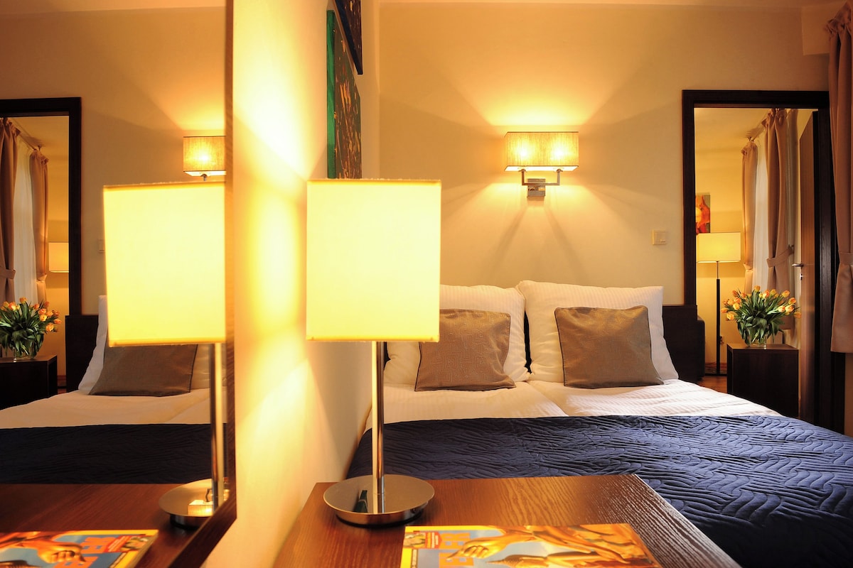 A hotel room with two beds, each with a bedside table and lamp, a large mirror, and a small table with a magazine.