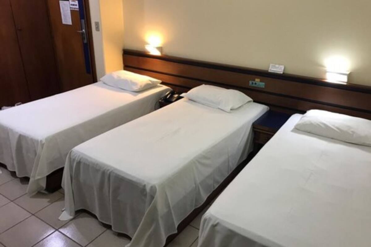 Une chambre d’hôtel avec trois lits simples, chacun doté de draps blancs et d’une tête de lit en bois. Une lampe est fixée au mur au-dessus des lits, et une porte se trouve sur le côté gauche.