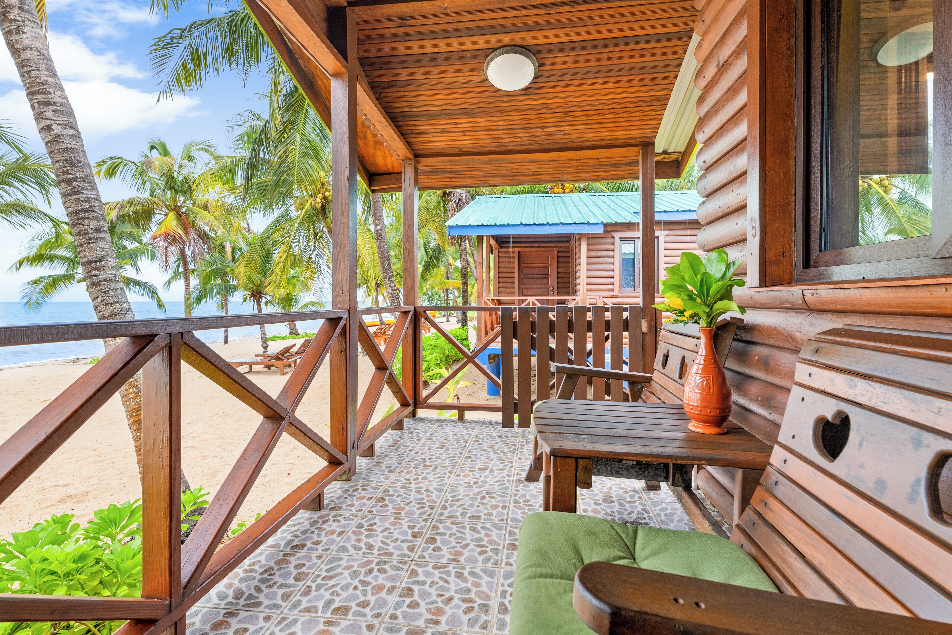 A wooden cabin with a balcony overlooking a beach, featuring a stone-tiled floor and wooden furniture.