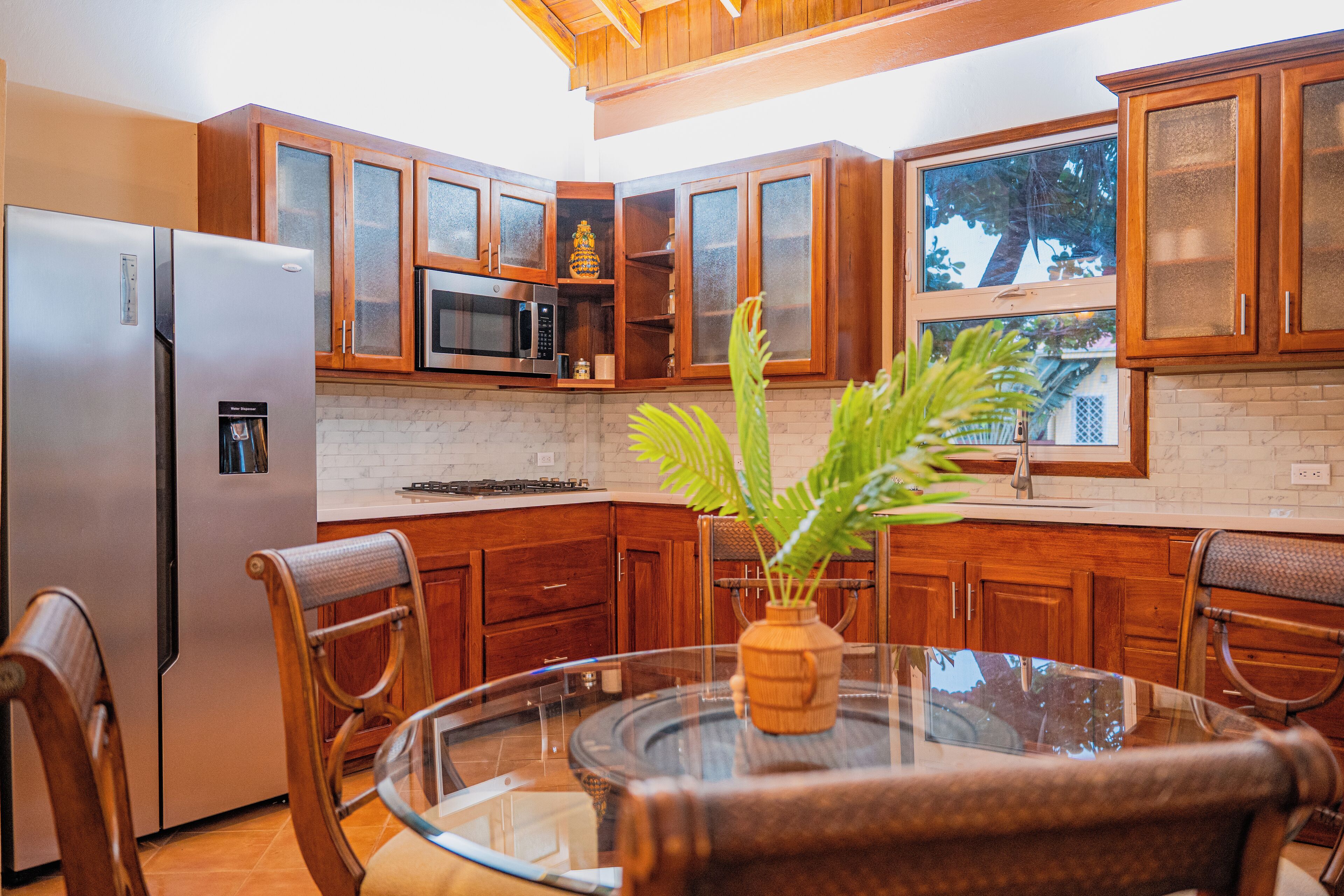 A kitchen with wooden cabinets, a stainless steel refrigerator, a microwave, and a glass dining table with chairs.