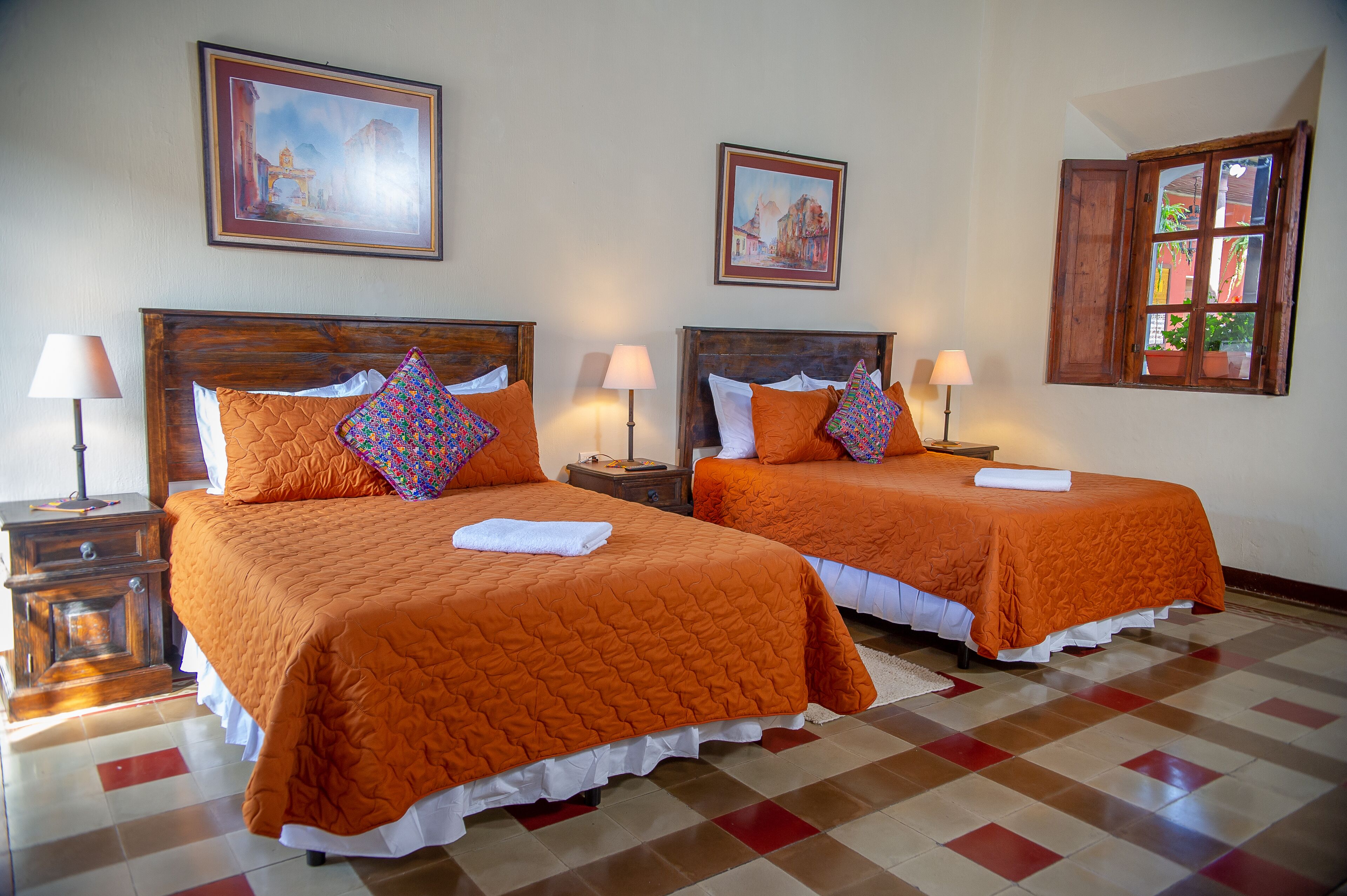Hotelkamer met twee bedden, elk voorzien van een oranje sprei en bijpassende kussens, een houten hoofdbord, een nachtkastje met een lamp en een raam met uitzicht op gebouwen.
