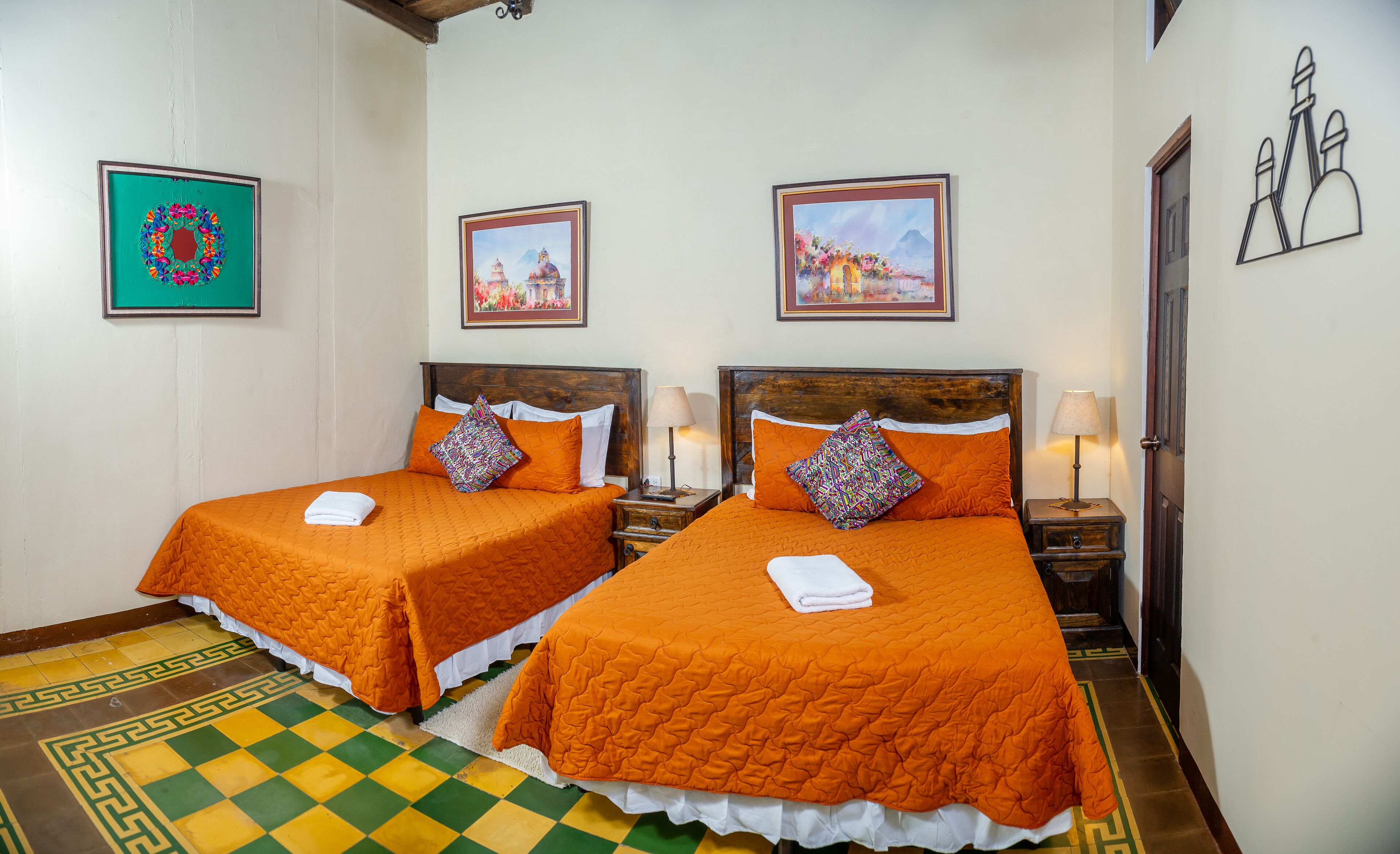 A hotel room with two beds, each with an orange bedspread and decorative pillows. There are framed pictures on the wall, a wooden headboard, and a checkered floor.