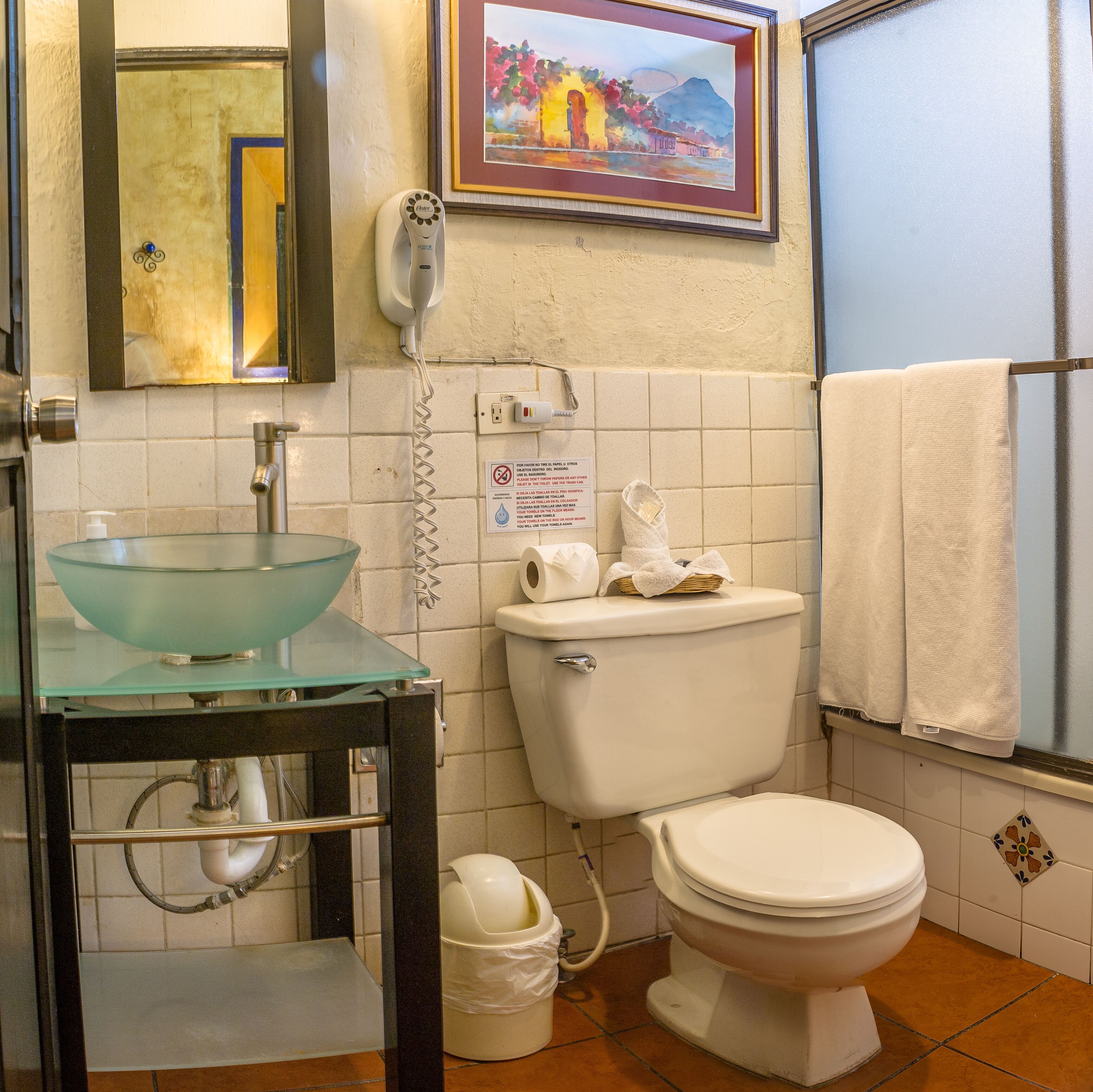 A bathroom with a glass sink, a toilet, a hand dryer, and a framed picture on the wall.