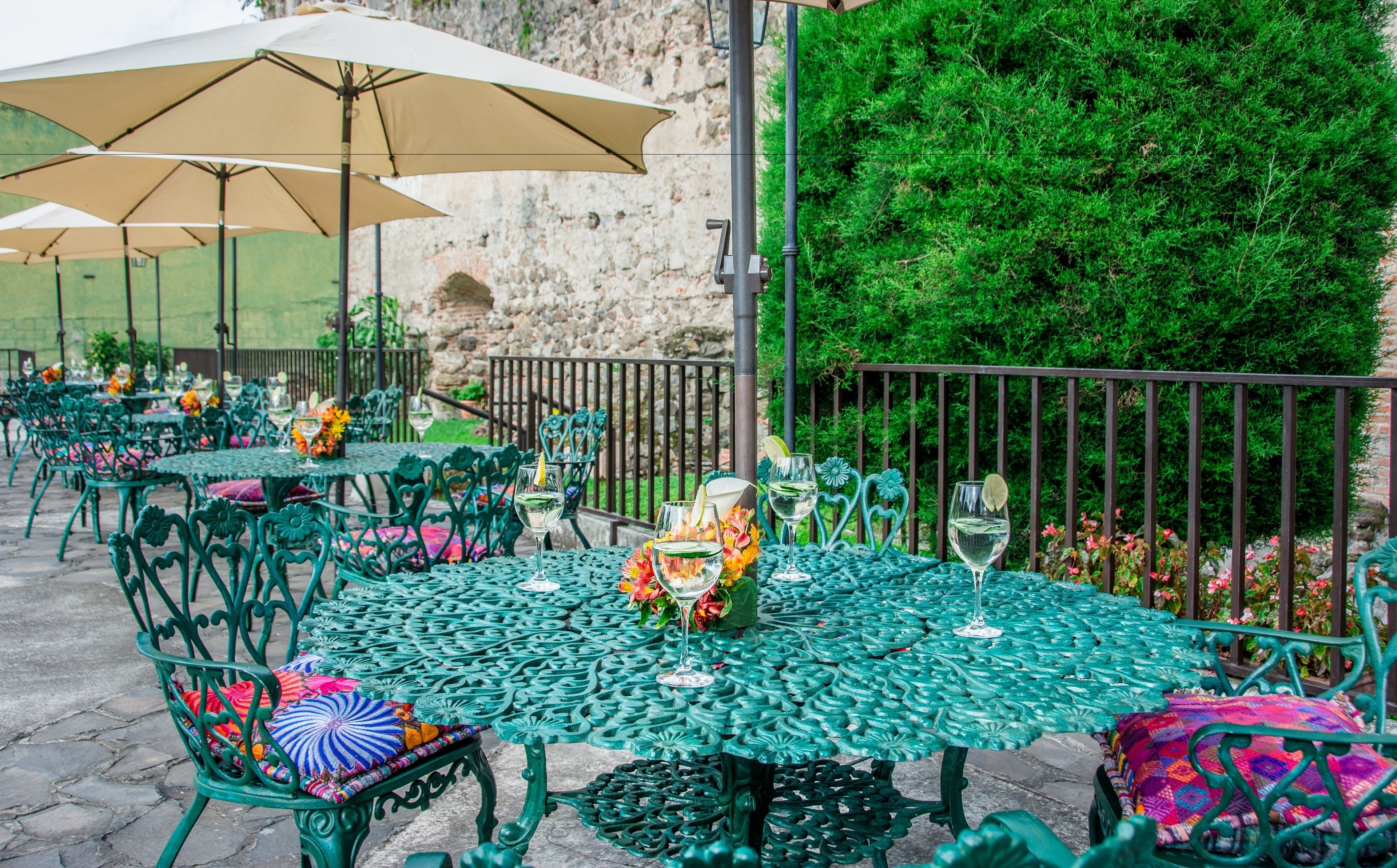 Espace extérieur aménagé avec des tables et des chaises vertes, des parasols et un mur de pierre en arrière-plan.