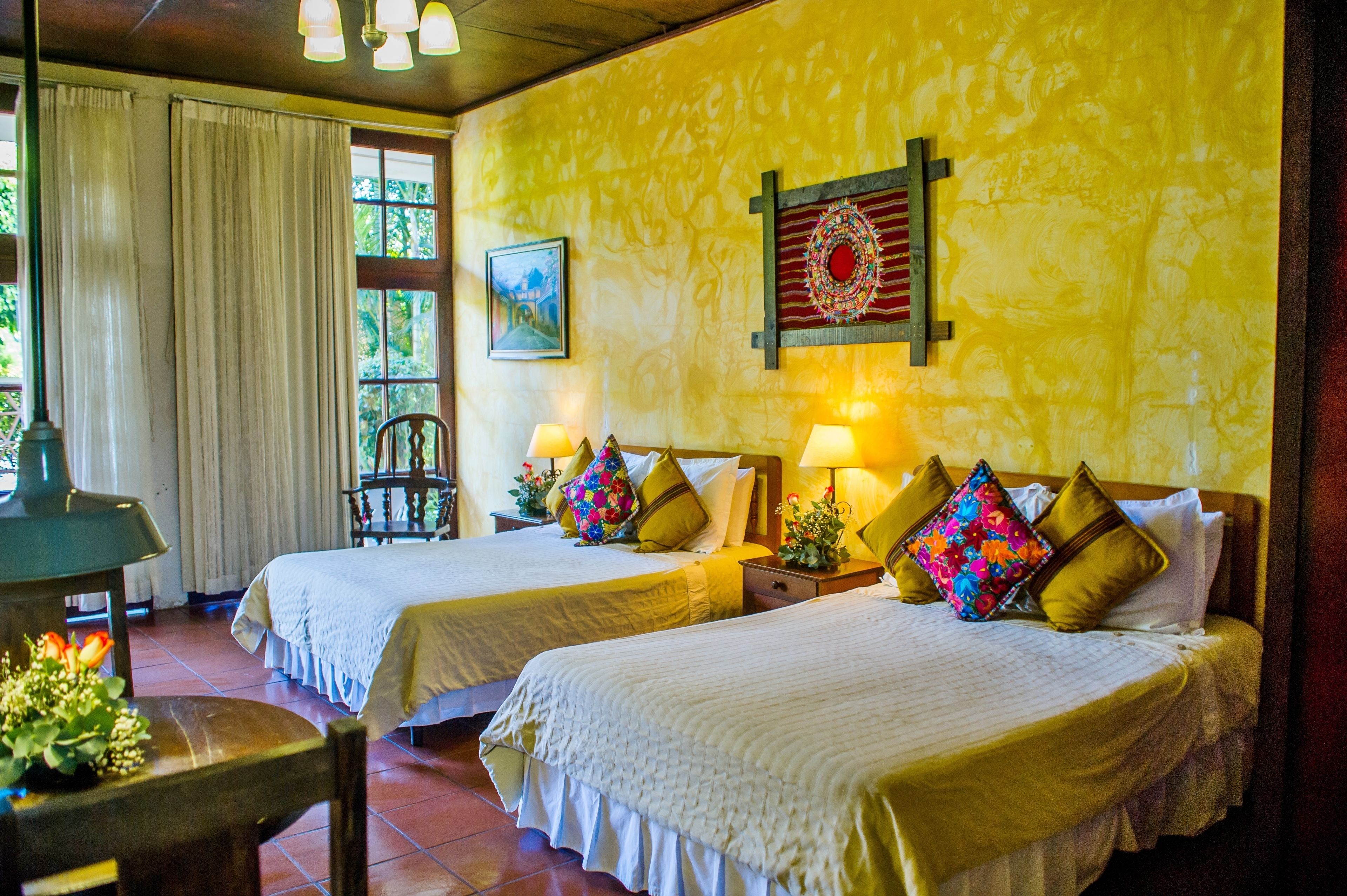 Une chambre avec deux lits, chacun doté d’une table de chevet et d’une lampe. On y trouve également une chaise et une table ornée de fleurs. La chambre est pourvue d’une fenêtre avec des rideaux et d’un tableau accroché au mur.