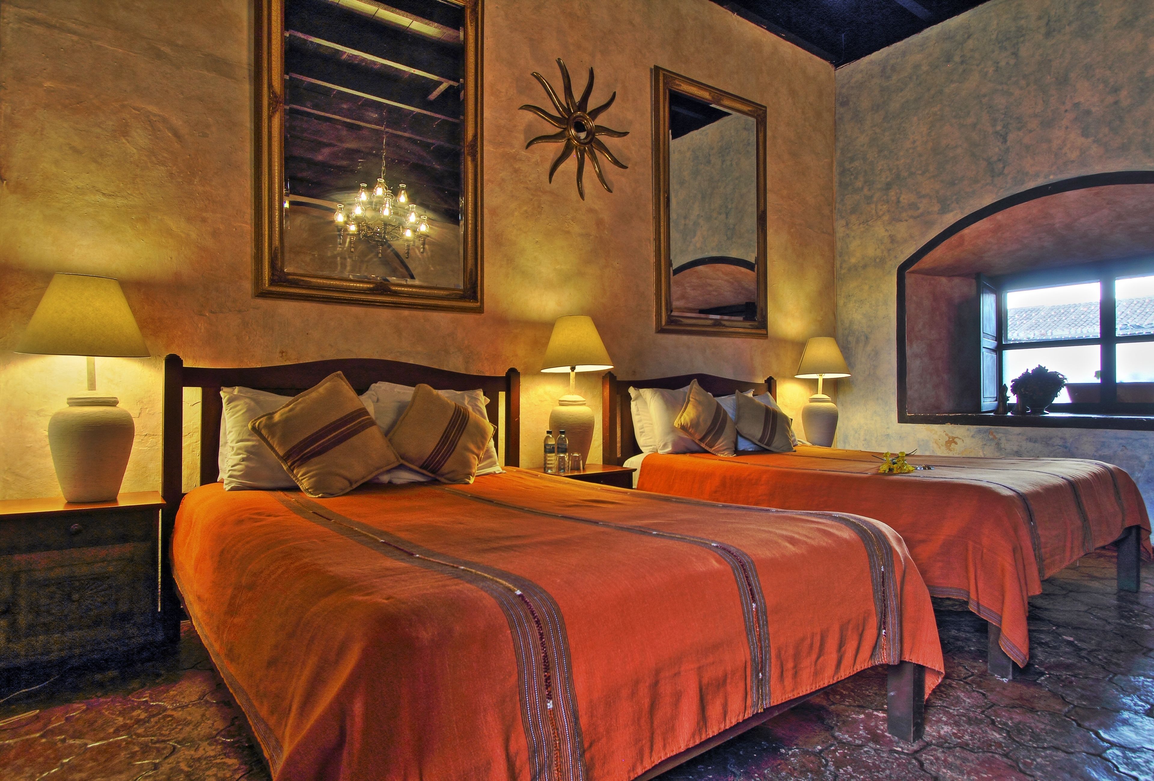 A hotel room with two beds, each with a wooden headboard, a large mirror, and a chandelier.