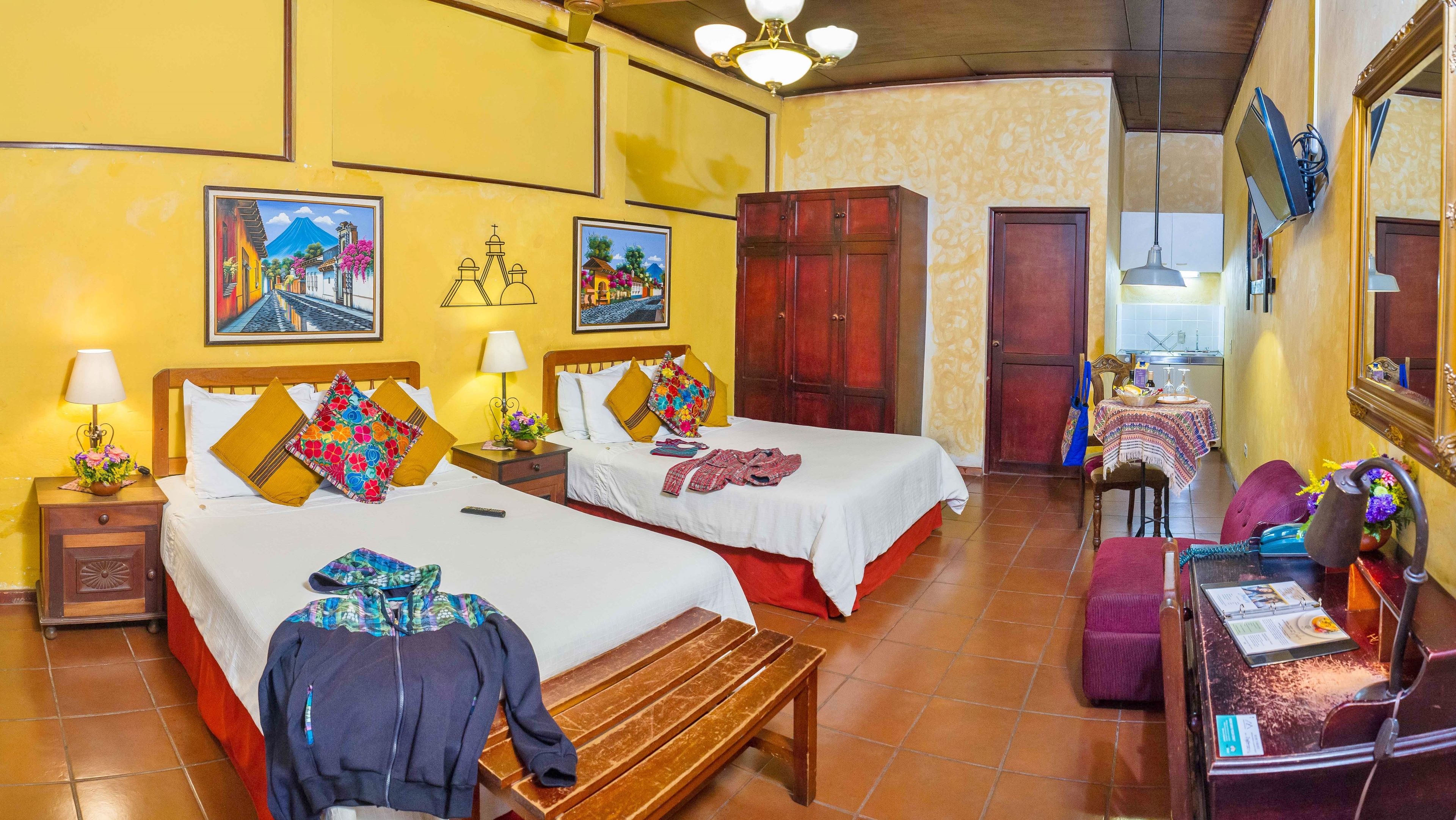 A hotel room with two beds, a wooden bench, a desk with a telephone, and a table with a teapot.
