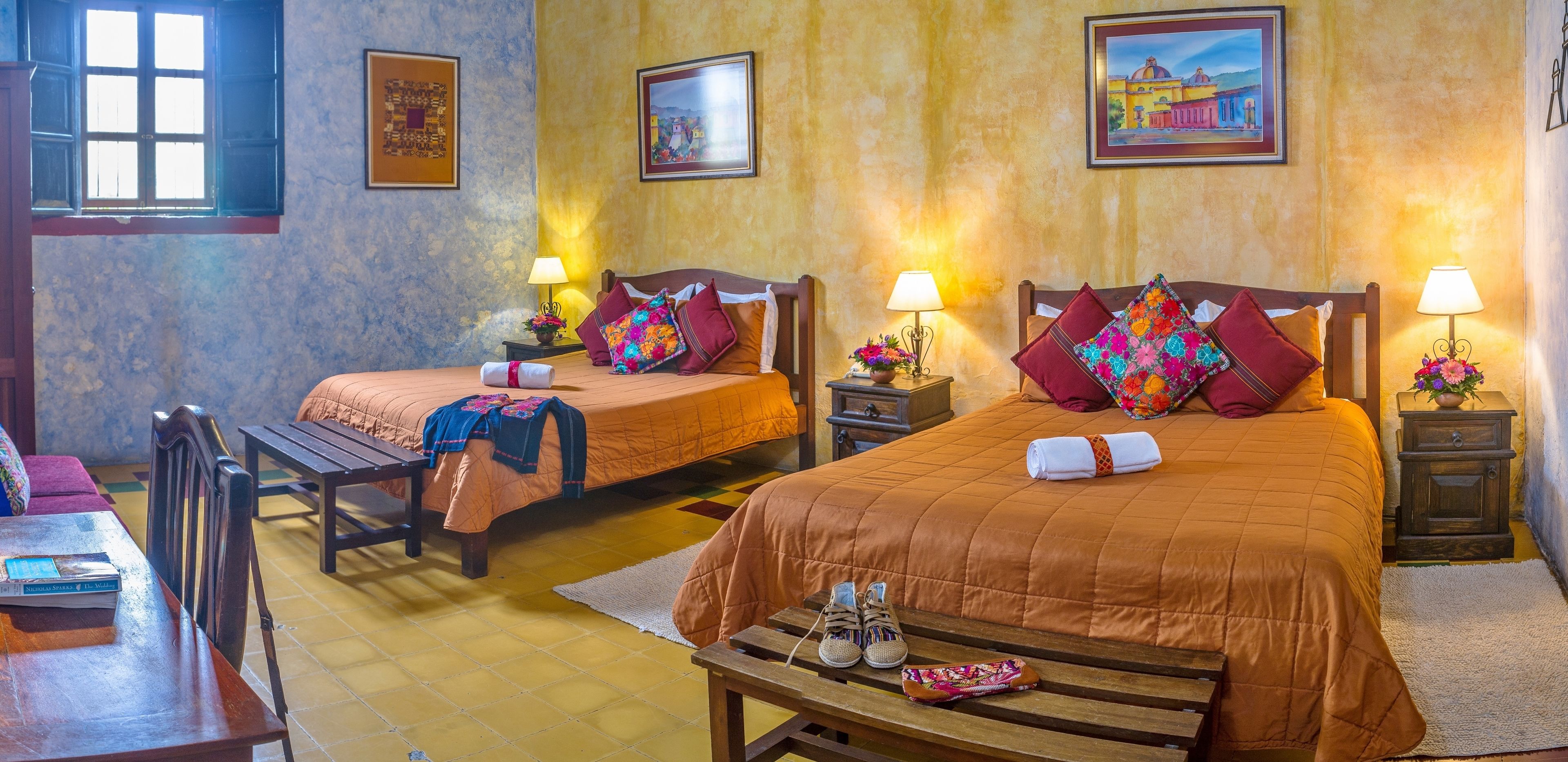 Habitación de hotel con dos camas, cada una con un almohadón y una lámpara. Hay un banco de madera con zapatos y un libro encima, y una mesita con un libro.