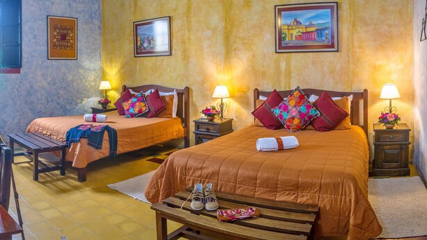 A hotel room with two beds, each with a pillow and a lamp. There is a wooden bench with shoes and a book on it, and a small table with a book. - Hotel Convento Santa Catalina (Antigua Guatemala)