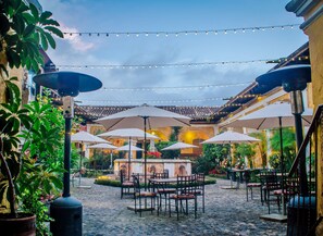 An outdoor patio area with tables and chairs under umbrellas, string lights, and potted plants. - Hotel Convento Santa Catalina (Antigua Guatemala)
