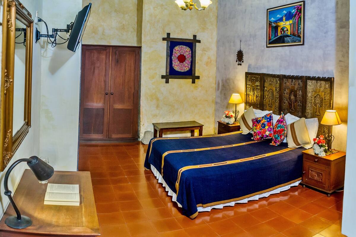 Una habitación de hotel con cama, mesitas de noche, escritorio, silla, armario y un cuadro en la pared.
