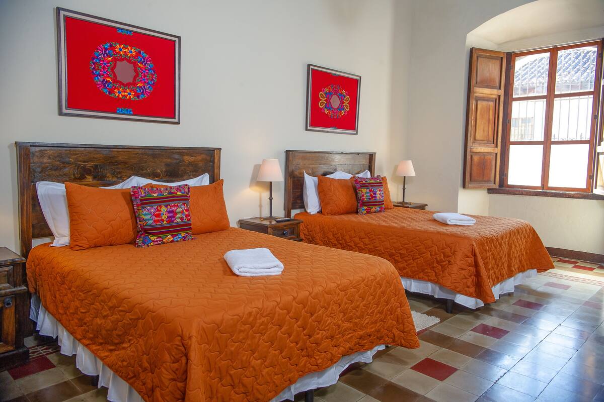 Habitación de hotel con dos camas, cada una con cubrecama naranja, almohadas blancas y una manta colorida. Hay dos cuadros enmarcados en la pared, una ventana con contraventanas de madera y un suelo de baldosas.