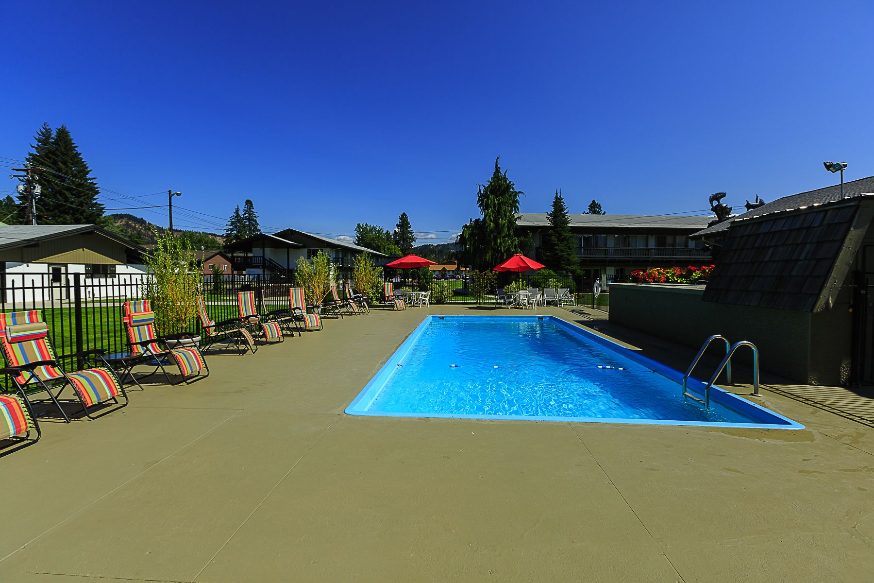 Una piscina con sillones de descanso y sombrillas, rodeada de edificios y árboles.