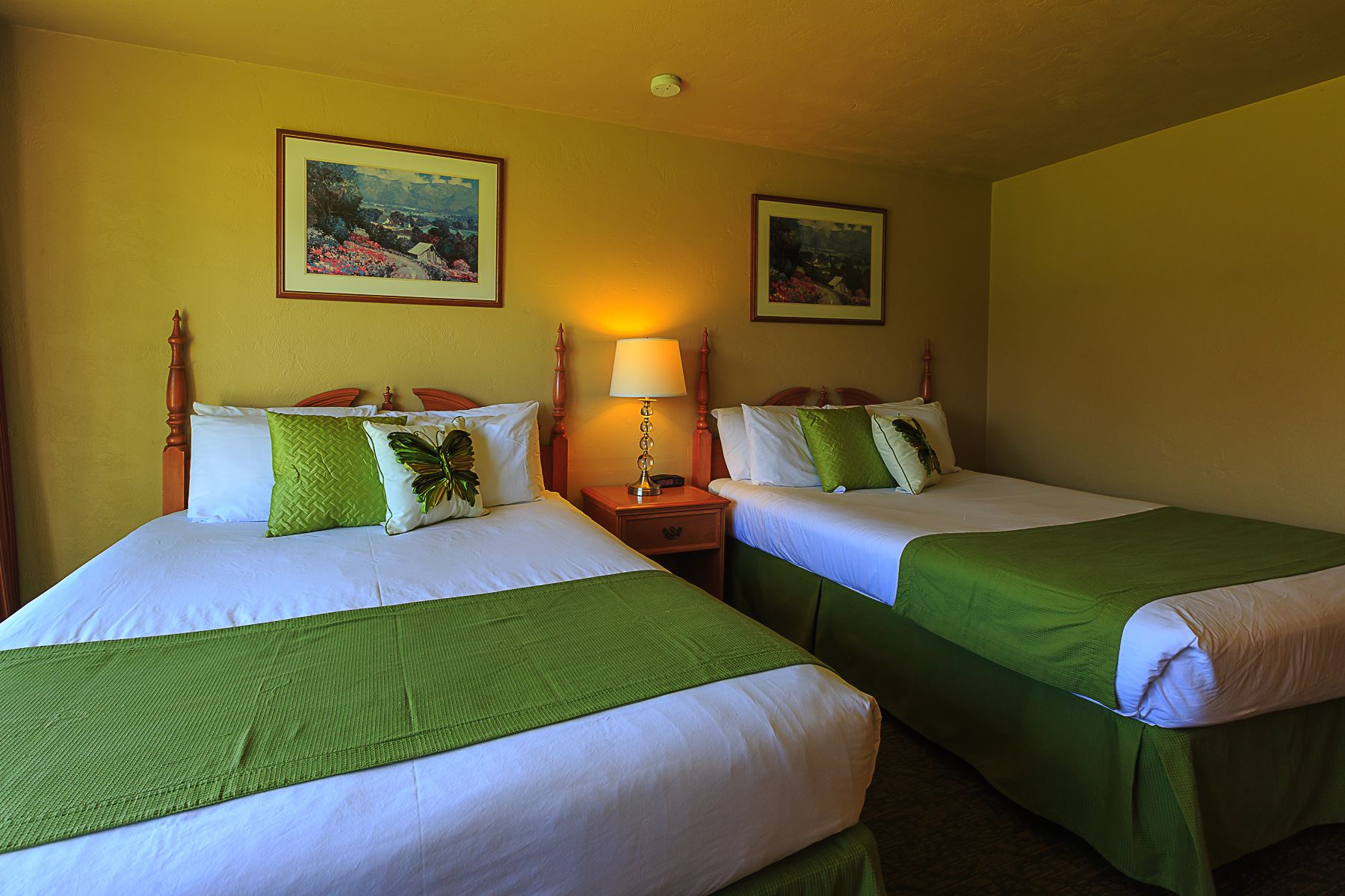 Une chambre d’hôtel avec deux lits, chacun recouvert d’une couvre-lit verte et doté de draps blancs. Deux tableaux ornent les murs, une lampe se trouve sur la table de chevet, et le lit situé à gauche est surmonté d’une tête de lit en bois.