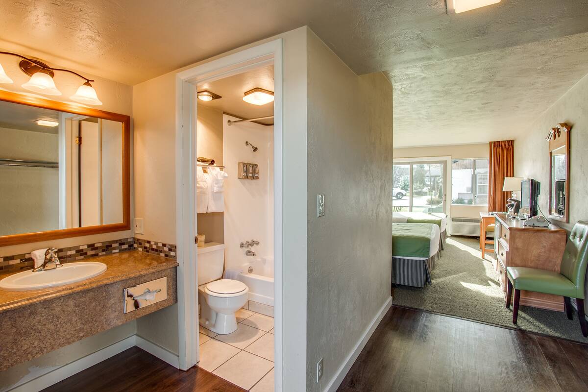 A bathroom with a sink, toilet, and shower. There is a mirror above the sink and a window in the adjacent room.