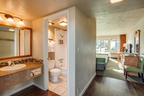 A bathroom with a sink, toilet, and shower. There is a mirror above the sink and a window in the adjacent room.
