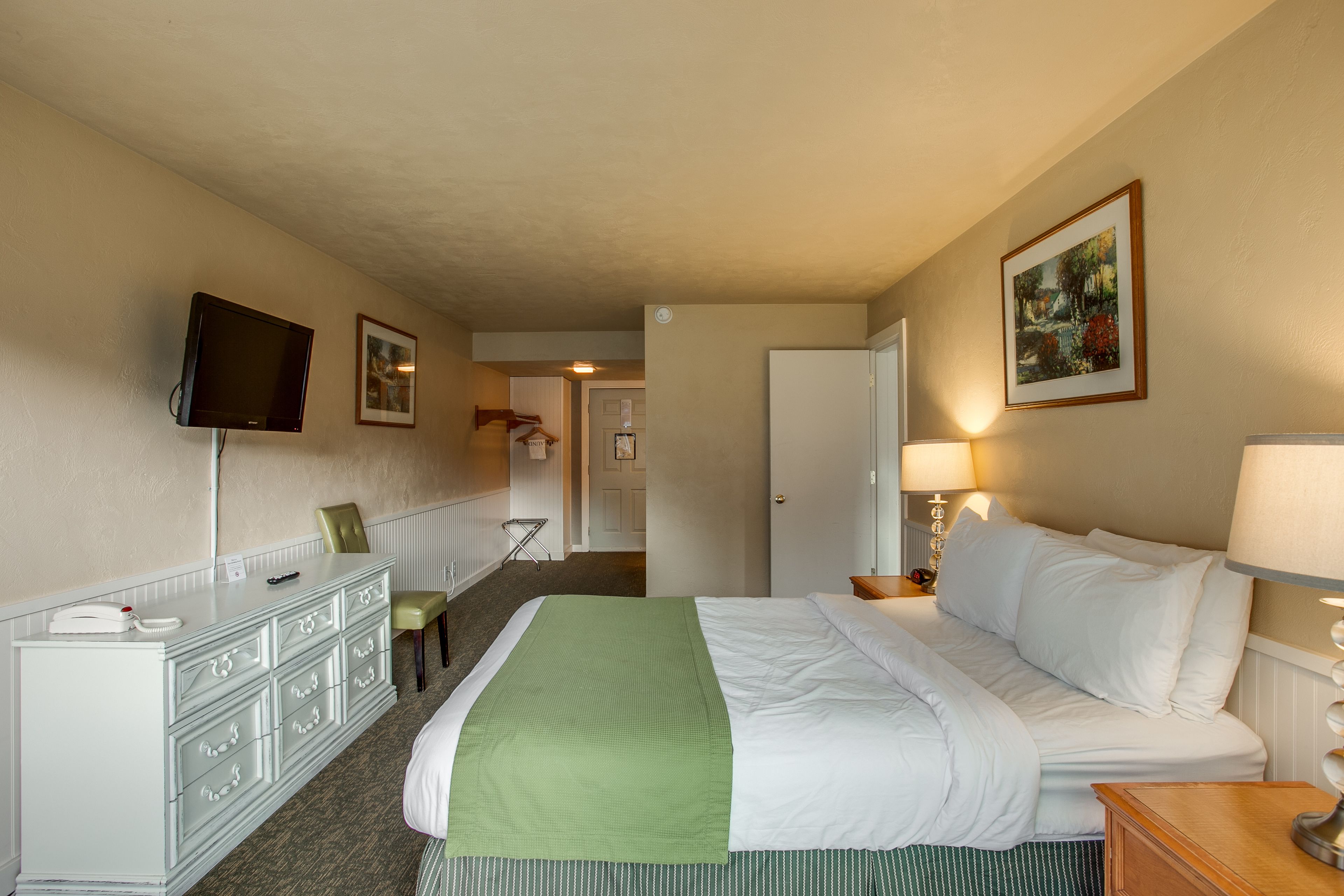 A hotel room with a bed, a dresser, a television mounted on the wall, and a door leading to another room.