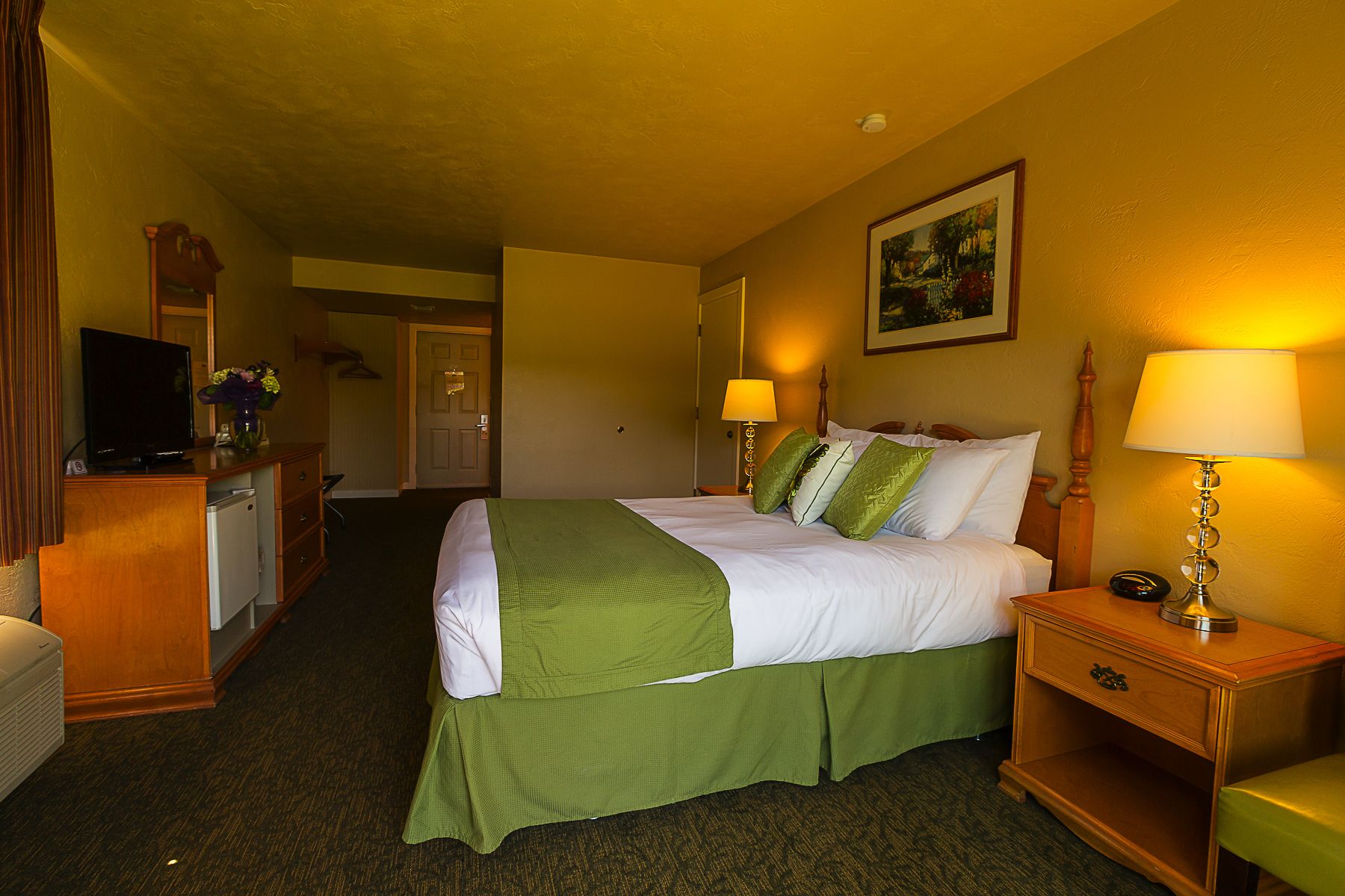 Una habitación de hotel con cama, lámparas de noche, televisión, cómoda y puerta que da a otra habitación.